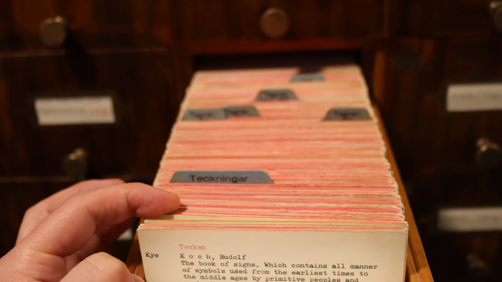 A hand pulling a single card from a library catalogue drawer: a queue indexed well enough that the next item can be taken without asking anybody