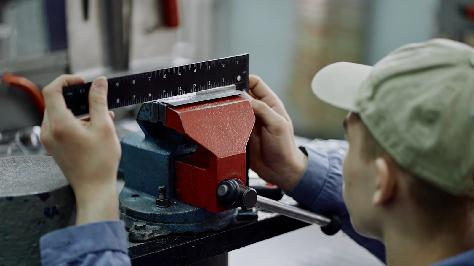 A worker measuring a metal part clamped in a vise, checking the finished thing against the figure it was supposed to hit rather than against an impression