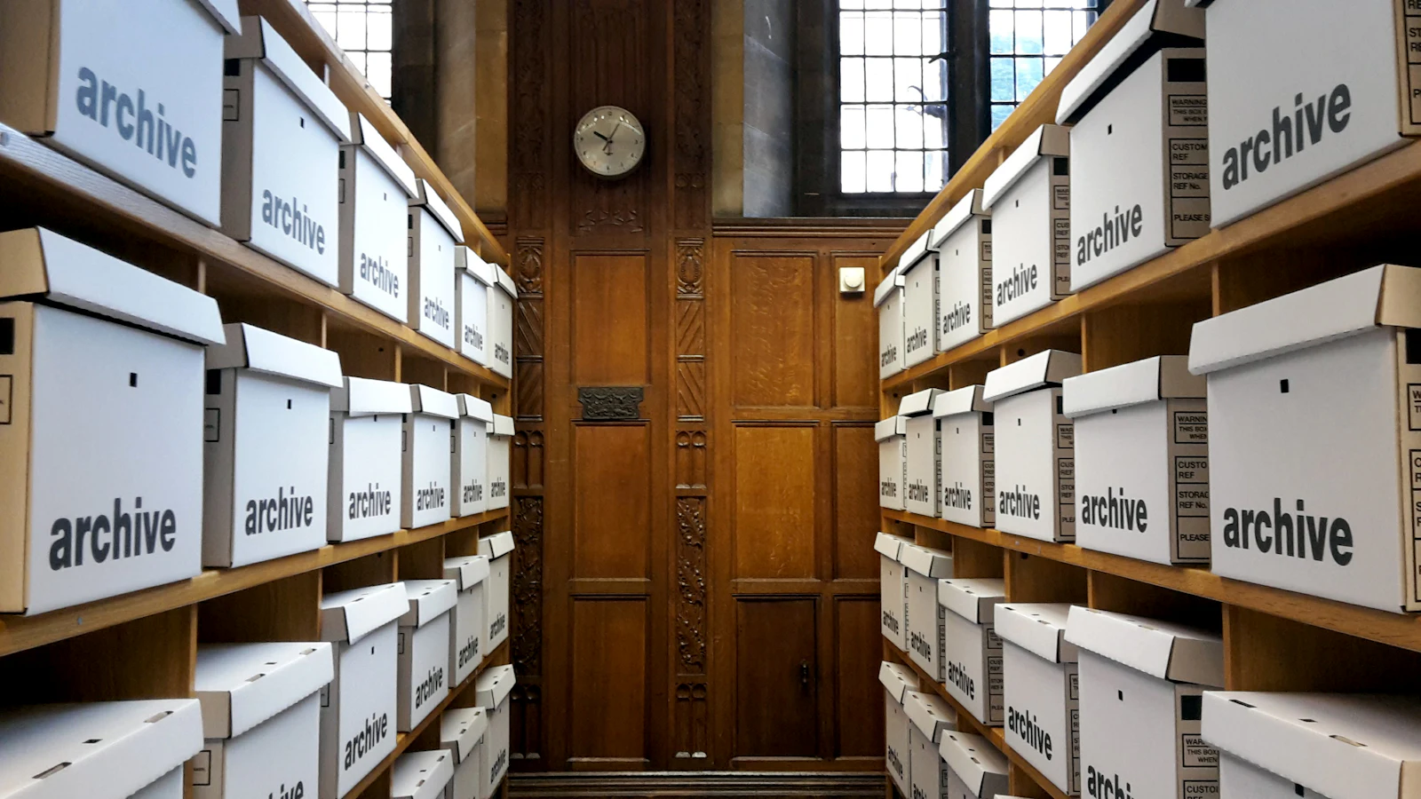 Two walls of identical boxes labelled only archive, receding to a panelled door and a clock, each saying what it is and nothing about what is inside it