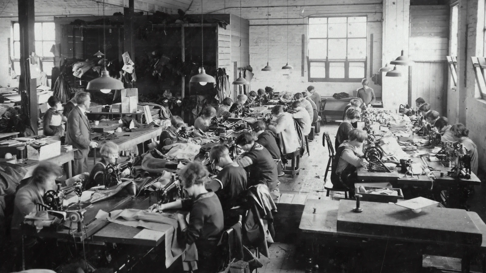 An archival photograph of a garment workshop: two long rows of sewing machines, every operator at their own station with their own work in front of them, and one supervisor standing over the floor