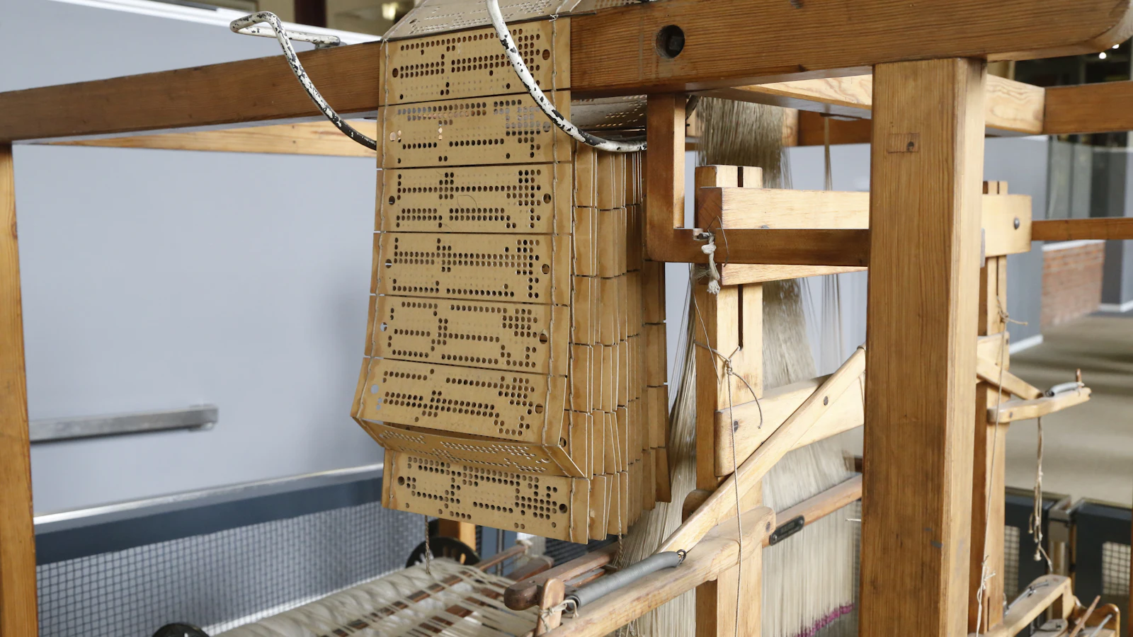 A chain of punched Jacquard cards laced together above a loom's warp threads, a pattern written in the form the machine reads directly rather than one an operator has to interpret first